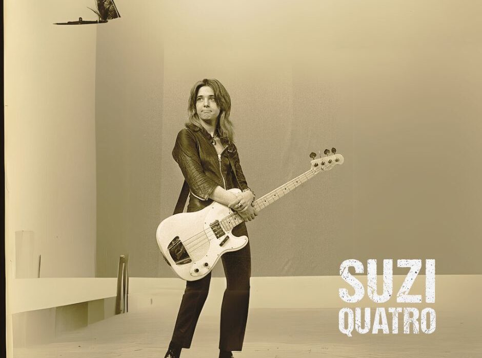 Suzi Quatro performing live on stage, wearing her iconic leather outfit and playing bass guitar.